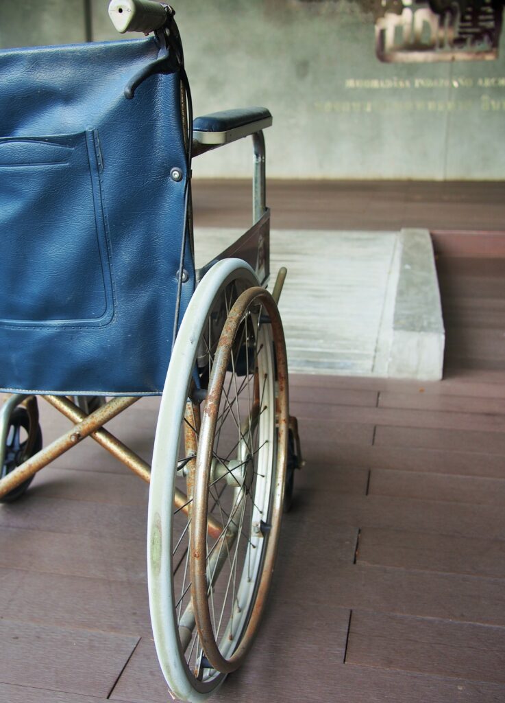 Close-up of an old wheelchair's large rear wheel with a rusted metal rim, blue vinyl seat back, and worn frame, positioned on a wooden deck near a ramp.