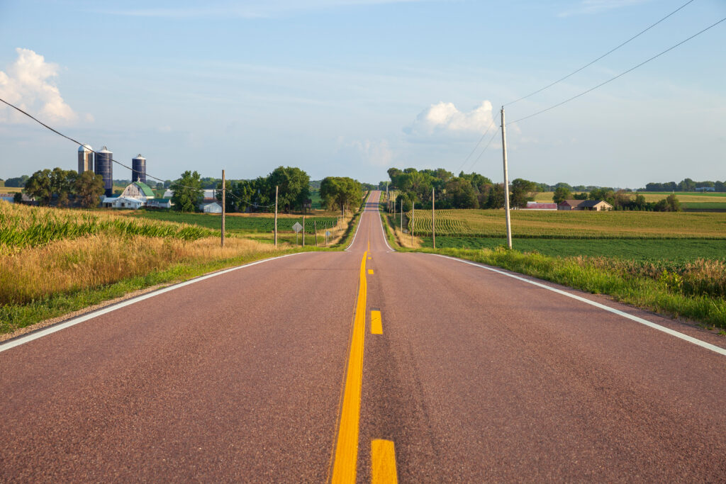 A straight paved road stretches through farmland toward the horizon, flanked by cornfields, with a barn and grain silos visible in the distance under a partly cloudy sky.