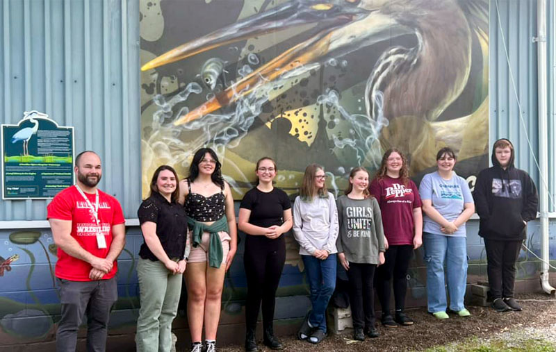 A group of nine people, including one adult man and several teenage girls and one teenage boy, pose smiling in front of a large mural of a heron catching a fish on the side of a metal building.
