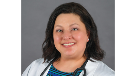 Professional headshot of a smiling woman with dark wavy hair, wearing a white lab coat over a blue patterned top, with a stethoscope around her neck, against a gray background.