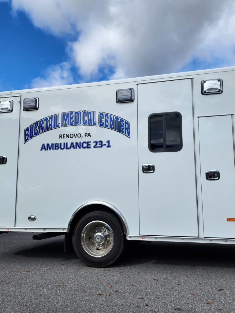 The side of a white ambulance labeled "Bucktail Medical Center, Renovo, PA, Ambulance 23-1" in blue lettering, parked under a partly cloudy sky.