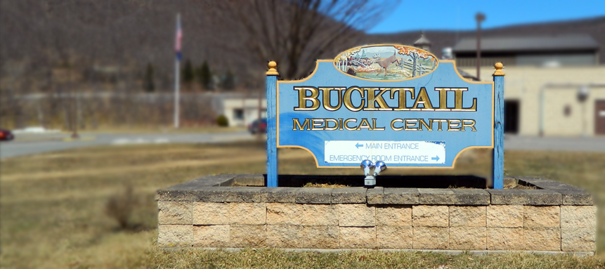 A blue and gold sign reading "Bucktail Medical Center" with directional arrows for the main entrance and emergency room entrance, topped with an oval image of a deer in a forest setting.