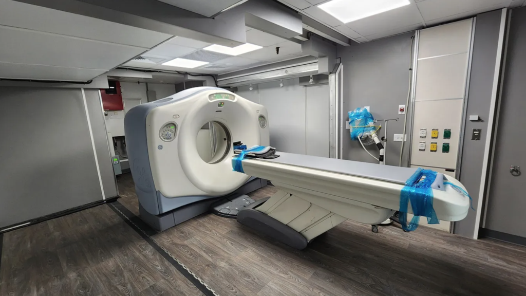 A white and blue CT scanner with a circular scanning tunnel and an extended patient table wrapped in blue protective plastic, situated in a gray-walled imaging room.