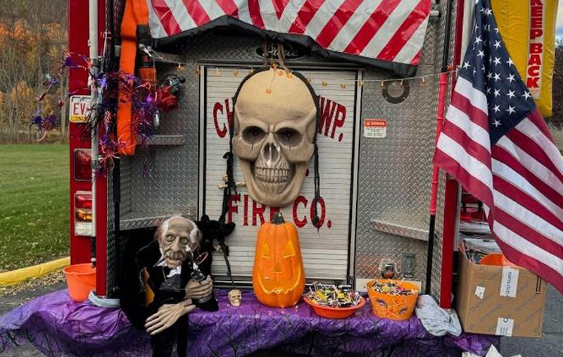 The back of a fire truck decorated for Halloween with a large skull, jack-o'-lantern, animatronic figure, spiderwebs, and candy bowls, alongside American flags.