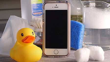 A smartphone in a case sits on a table surrounded by cleaning supplies, including a bottle of alcohol, cotton swabs, a blue sponge, cotton balls, a paper towel, and a rubber duck.