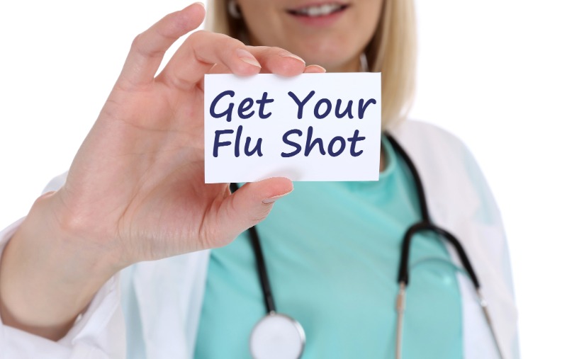 A smiling doctor wearing a white coat and stethoscope over teal scrubs holds up a card that reads "Get Your Flu Shot."