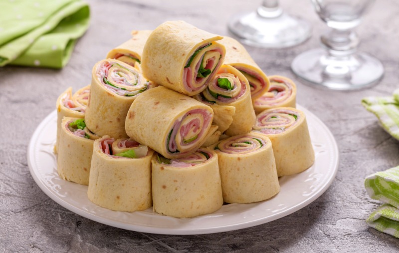 A white plate stacked with sliced tortilla pinwheel sandwiches filled with ham, cheese, and lettuce, arranged in a pyramid shape.