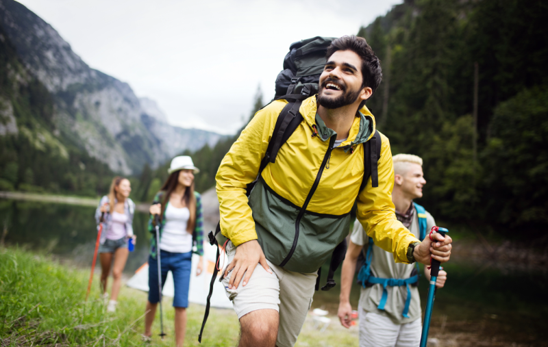 A group of four hikers with backpacks and trekking poles smile and laugh while walking near a lake surrounded by mountains and forest.
