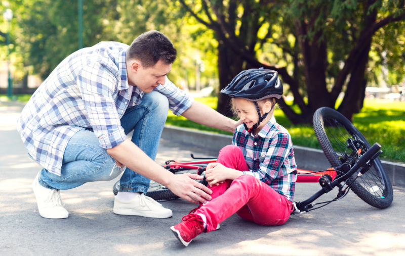 A man kneels to comfort a young child wearing a bike helmet who is sitting on the ground next to a fallen bicycle, holding their knee and crying, in a sunny park setting.