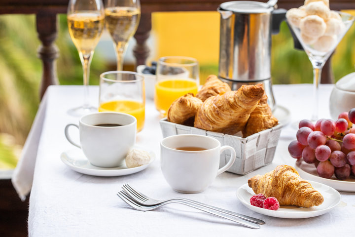 An elegant breakfast table setting with croissants in a basket, cups of coffee and tea, glasses of orange juice and champagne, grapes, and a small plate of raspberries and pastry.