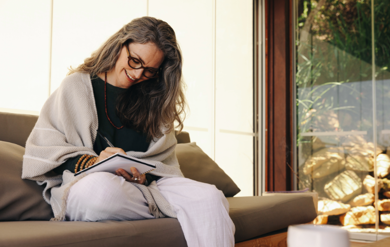 A woman with long gray hair and glasses, wrapped in a shawl, sits on a couch smiling while writing in a notebook, with a sunlit rock garden visible through a window behind her.