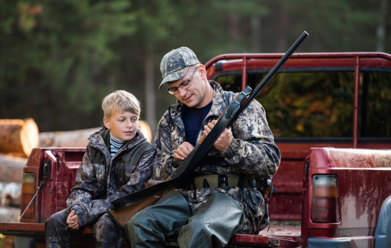 A man in camouflage hunting gear and a cap shows a rifle to a young boy, both seated on the tailgate of a red pickup truck near a forest.