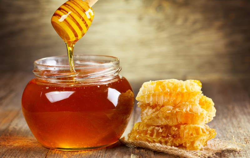 A glass jar of honey with a wooden dipper drizzling honey into it, next to a stack of golden honeycomb pieces resting on a burlap cloth.