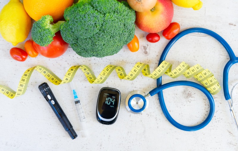 A flat lay of diabetes management tools including a lancing device, syringe, blood glucose meter reading "104," and a blue stethoscope, surrounded by fresh vegetables and fruit and a yellow measuring tape.