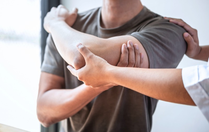 A physical therapist's hands support and examine a patient's bent arm and shoulder, with the patient wearing a brown t-shirt.