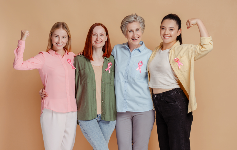 Four smiling women of different ages, each wearing a pink breast cancer awareness ribbon, standing arm-in-arm with two of them flexing their biceps.