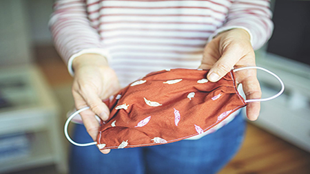 A person wearing a striped shirt and jeans holds up a reddish-orange cloth face mask with a small leaf pattern, using its elastic ear loops.