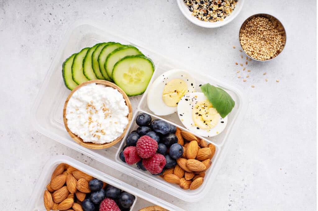 An overhead view of a compartmentalized bento-style lunch box filled with cucumber slices, cottage cheese, hard-boiled eggs, berries, and almonds, next to small bowls of seeds.